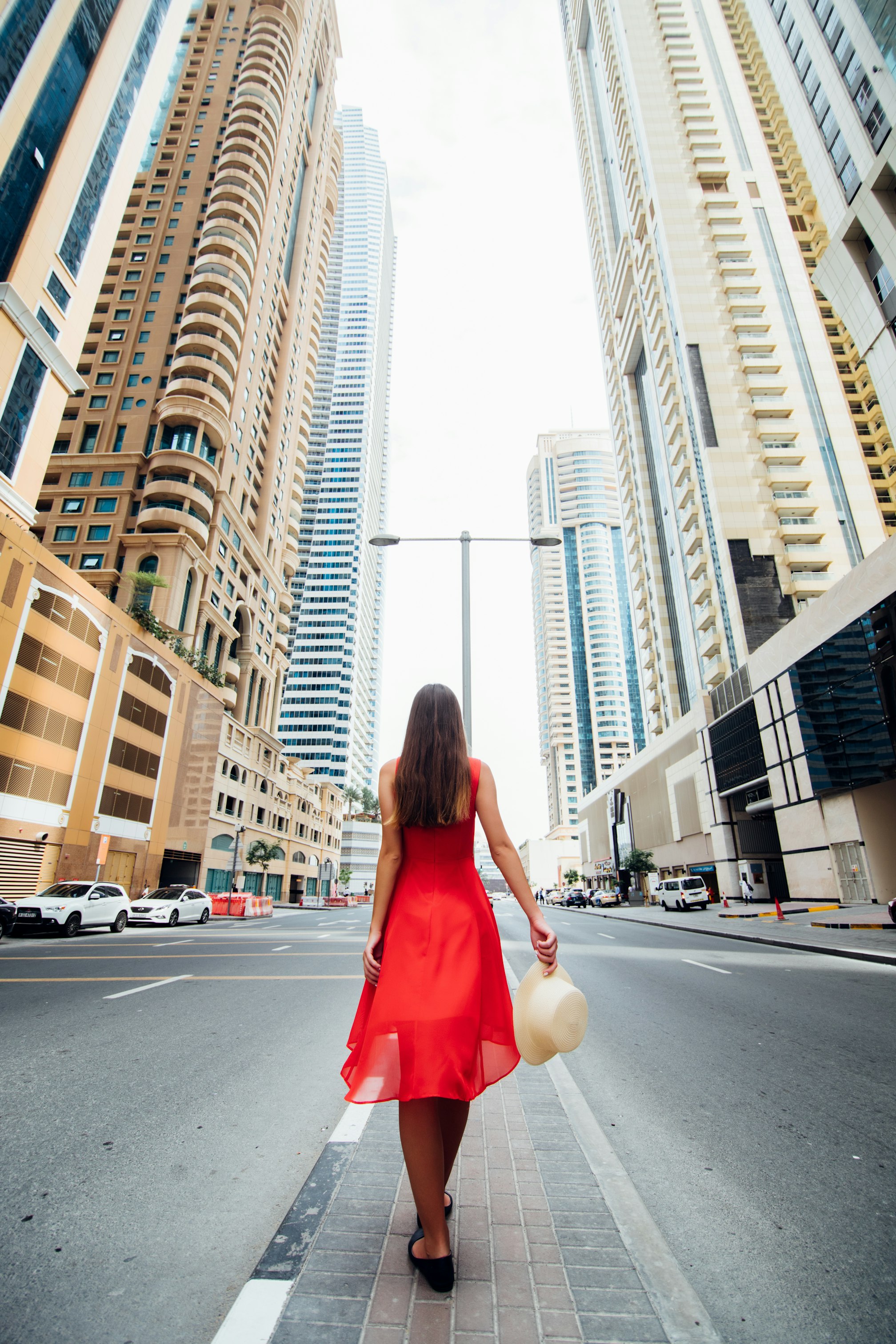 Young pretty woman in red dress and straw hat walking beetween skycrapers in modern city. Low angle view