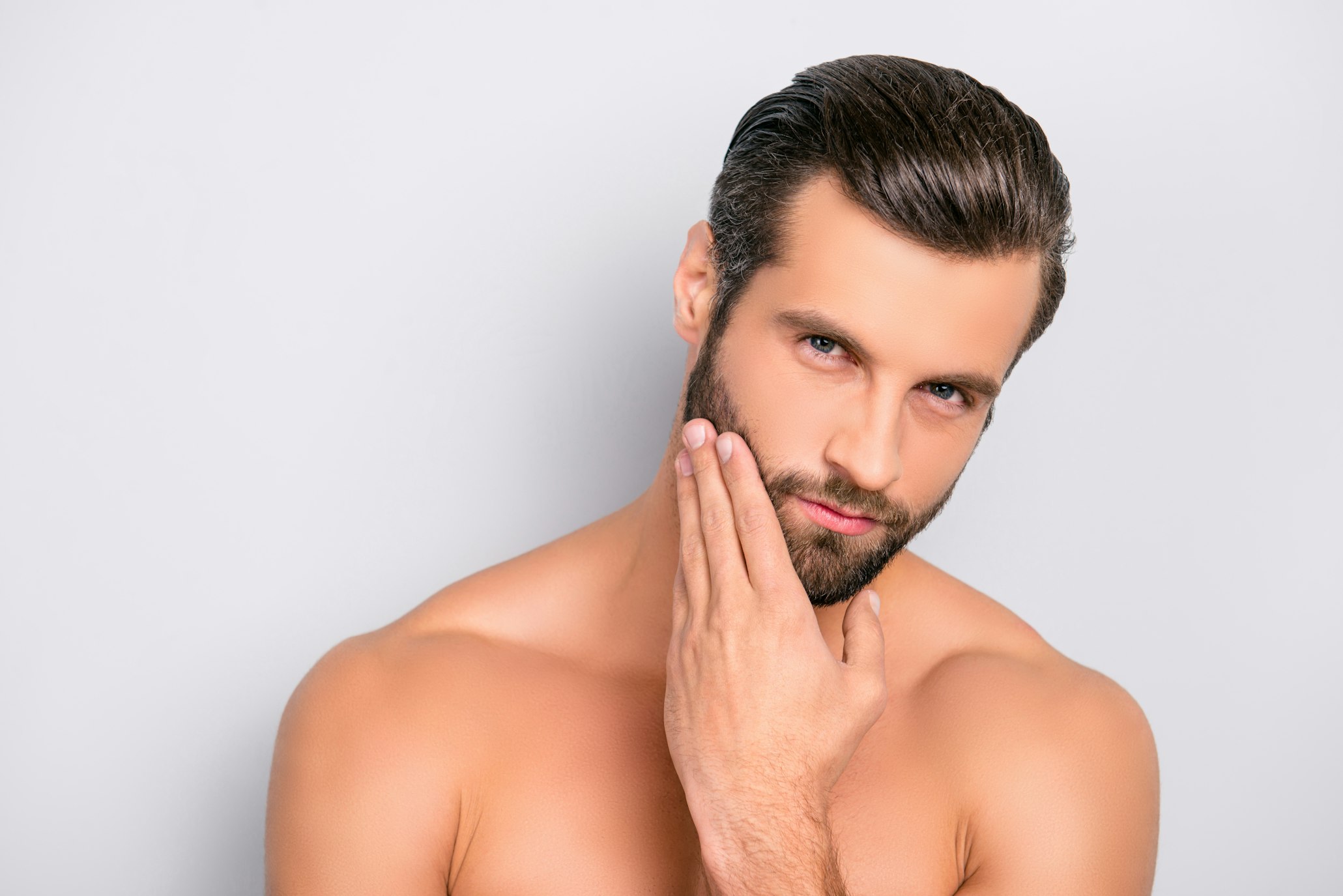 Attractive, brutal, modern, manly, virile, confident, dreamy, naked man touching his perfect, ideal face skin, holding hand on beard, cheek, looking at camera, isolated over gray