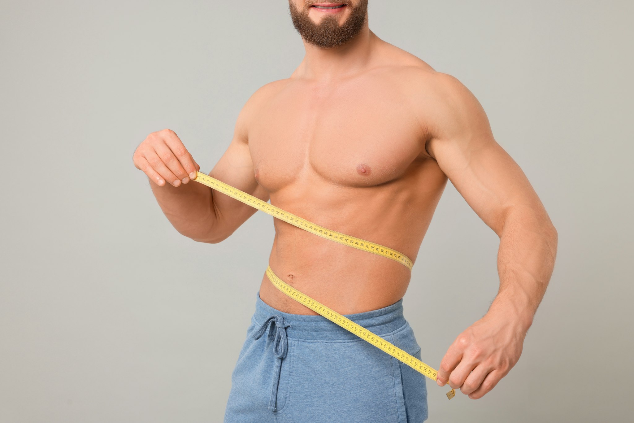 Athletic man measuring waist with tape on light grey background, closeup. Weight loss concept