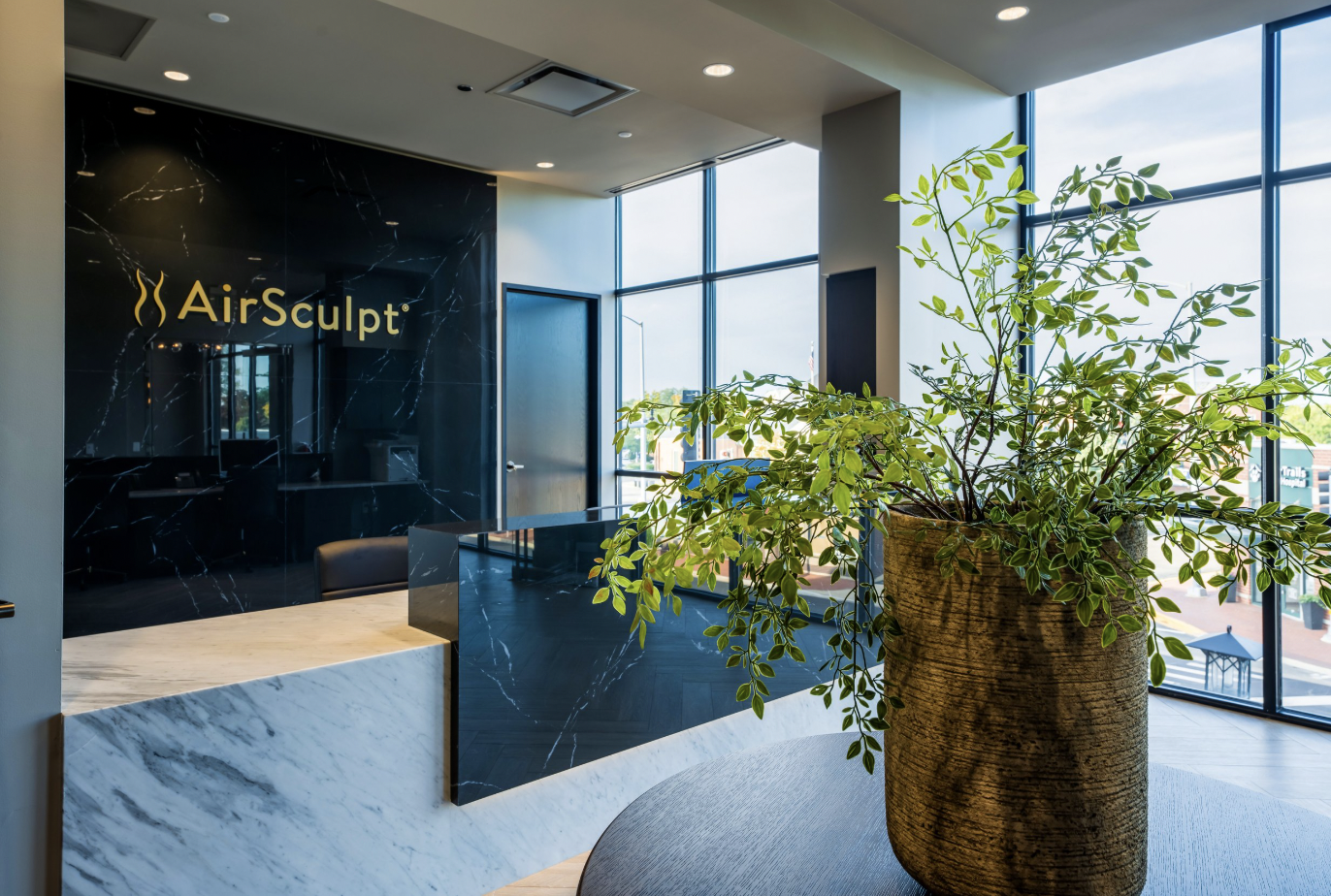 AirSculpt clinic lobby - plant in the foreground with black marble wall and golden AirSculpt logo