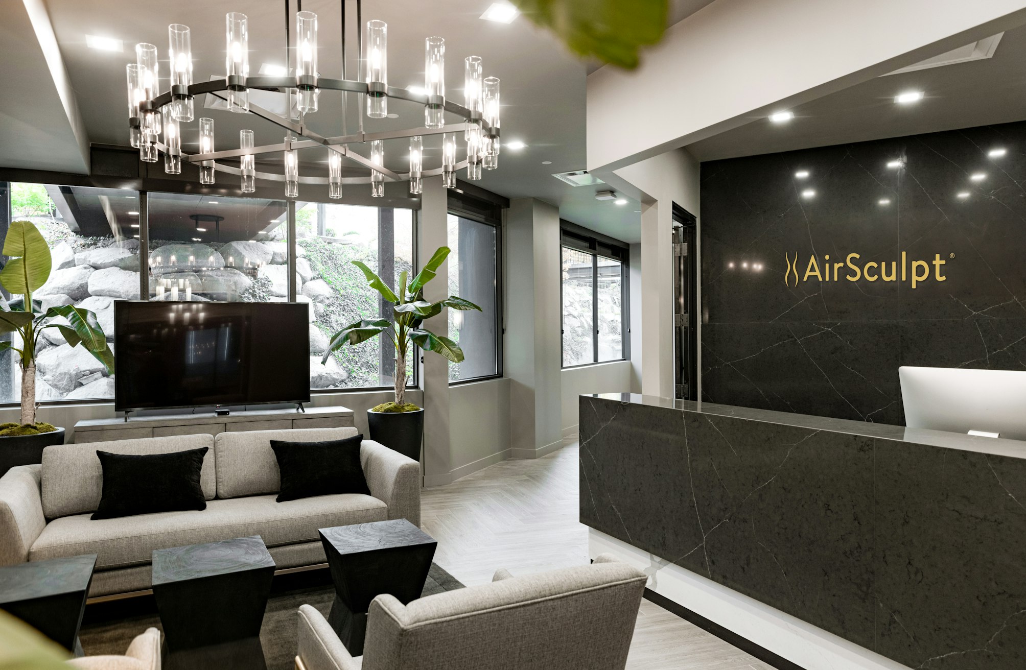 Lobby in AirSculpt Body Contouring Salt Lake City clinic with couch, wall‑mounted television, potted plants, chandelier and backdrop featuring AirSculpt logo