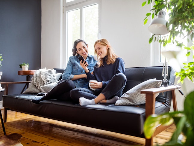 Happy young female friends sitting together on couch looking at a mobile phone