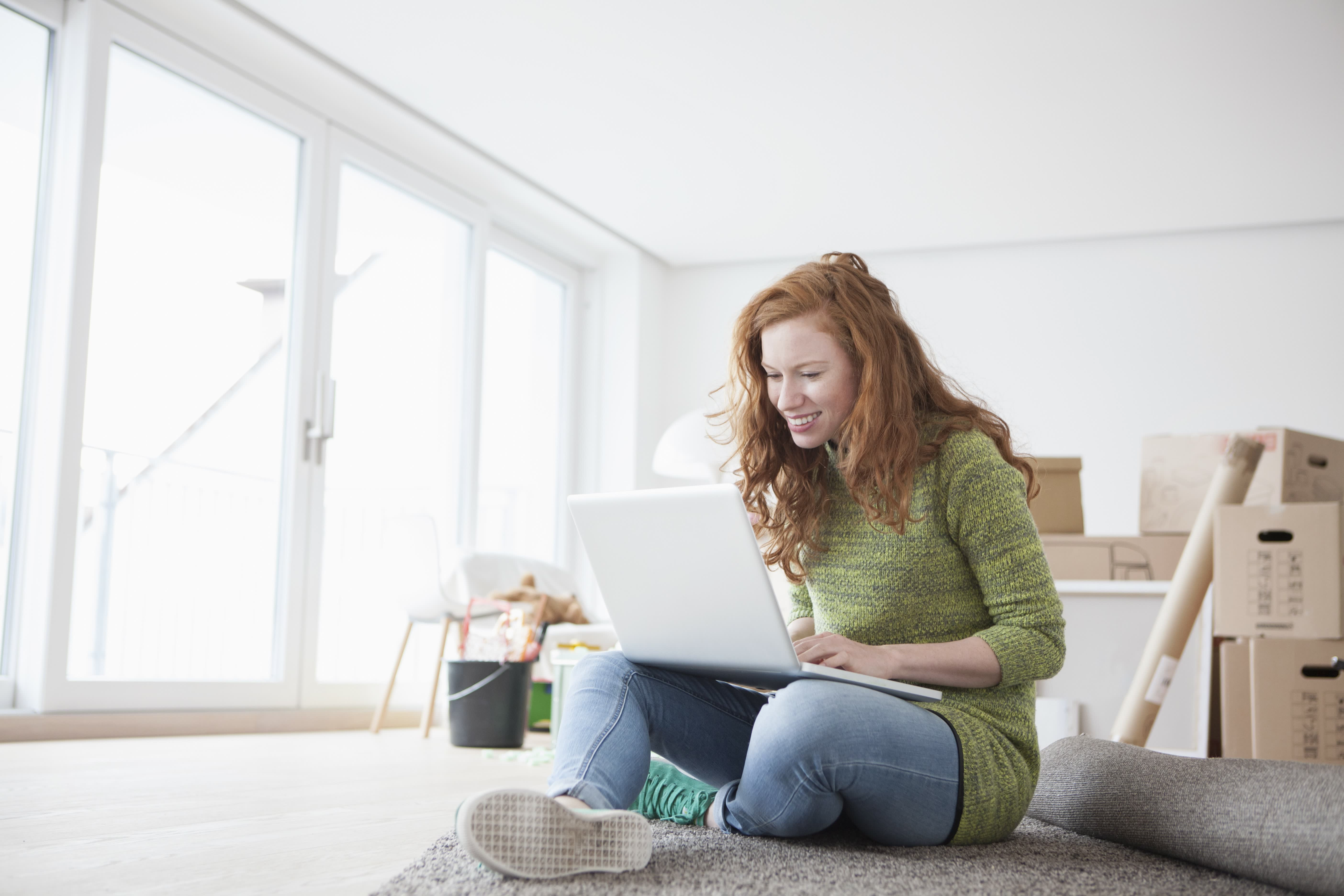 Young woman in new flat with cardboard boxes using laptop