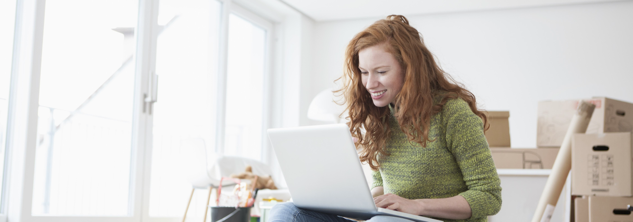 Young woman in new flat with cardboard boxes using laptop