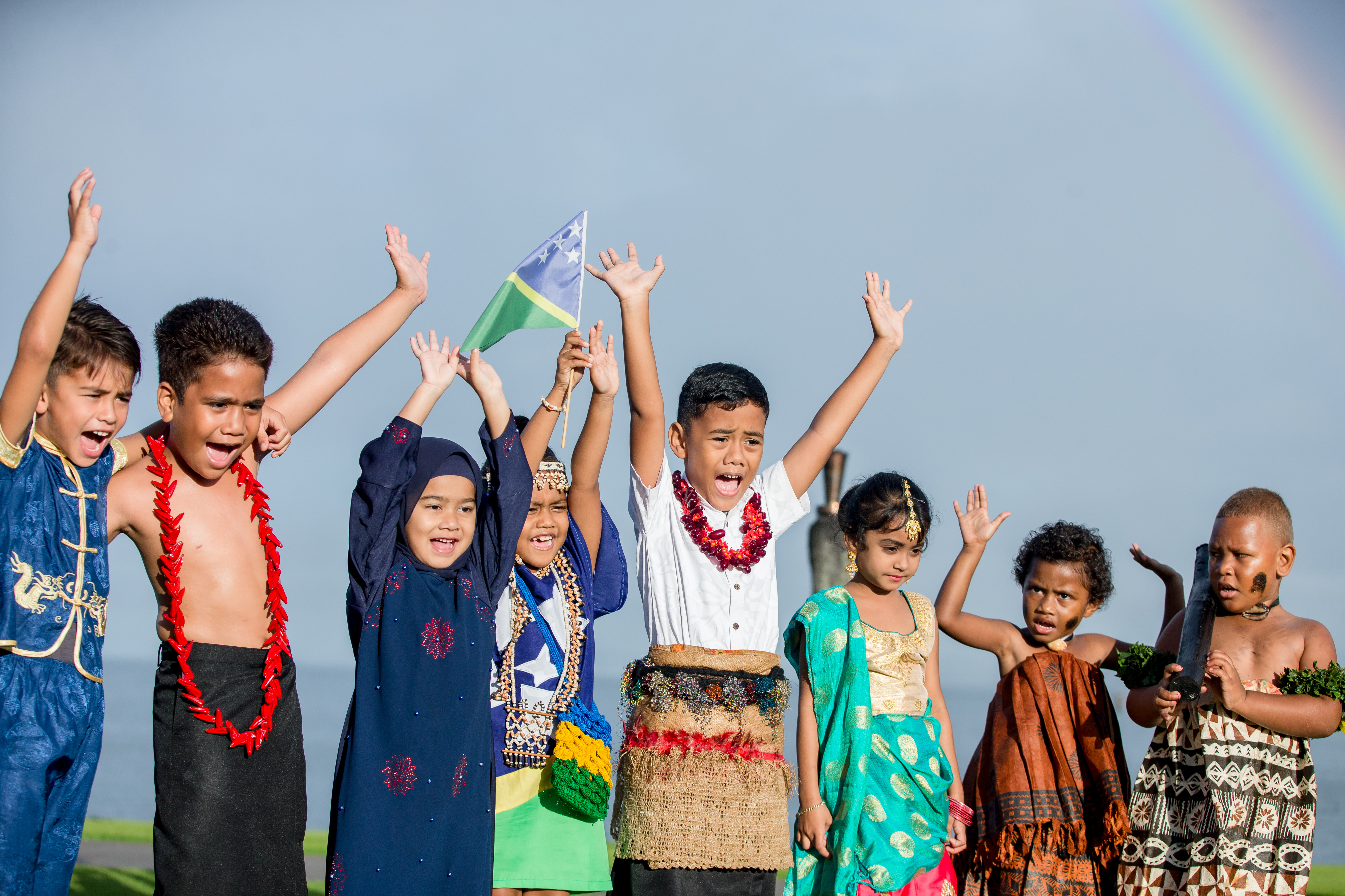 Young children stand together, raising their hands in a celebratory mood, dressed in their Pacific traditional attire, with a rainbow in the sky behind them. 