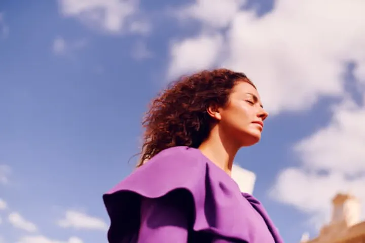 Mujer de piel blanca, pelo rizado y rojizo que fluye hacia atrás por el viento,  vistiendo una blusa morada. Tiene los ojos cerrados, de perfil. Al fondo, cielo azul y nubes.