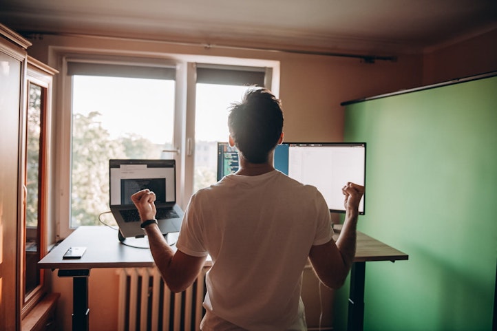 Kuba cieszy się z projektu strony internetowej Kuba odwrócony plecami zaciska pięści ze szczęścia po rozwiązaniu wymagającego problemu. W Tle stanowisko komputerowe.