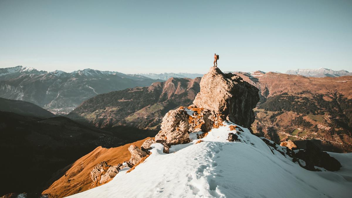 Ein Bergsteiger steht auf einem grossen Felsen in den schneebedeckten Bergen und blickt auf die Landschaft.