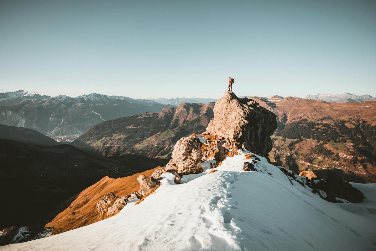 Ein Bergsteiger steht auf einem grossen Felsen in den schneebedeckten Bergen und blickt auf die Landschaft.