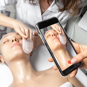 A hand holding smartphone making online live video stream of cosmetician doctor facial care procedure 