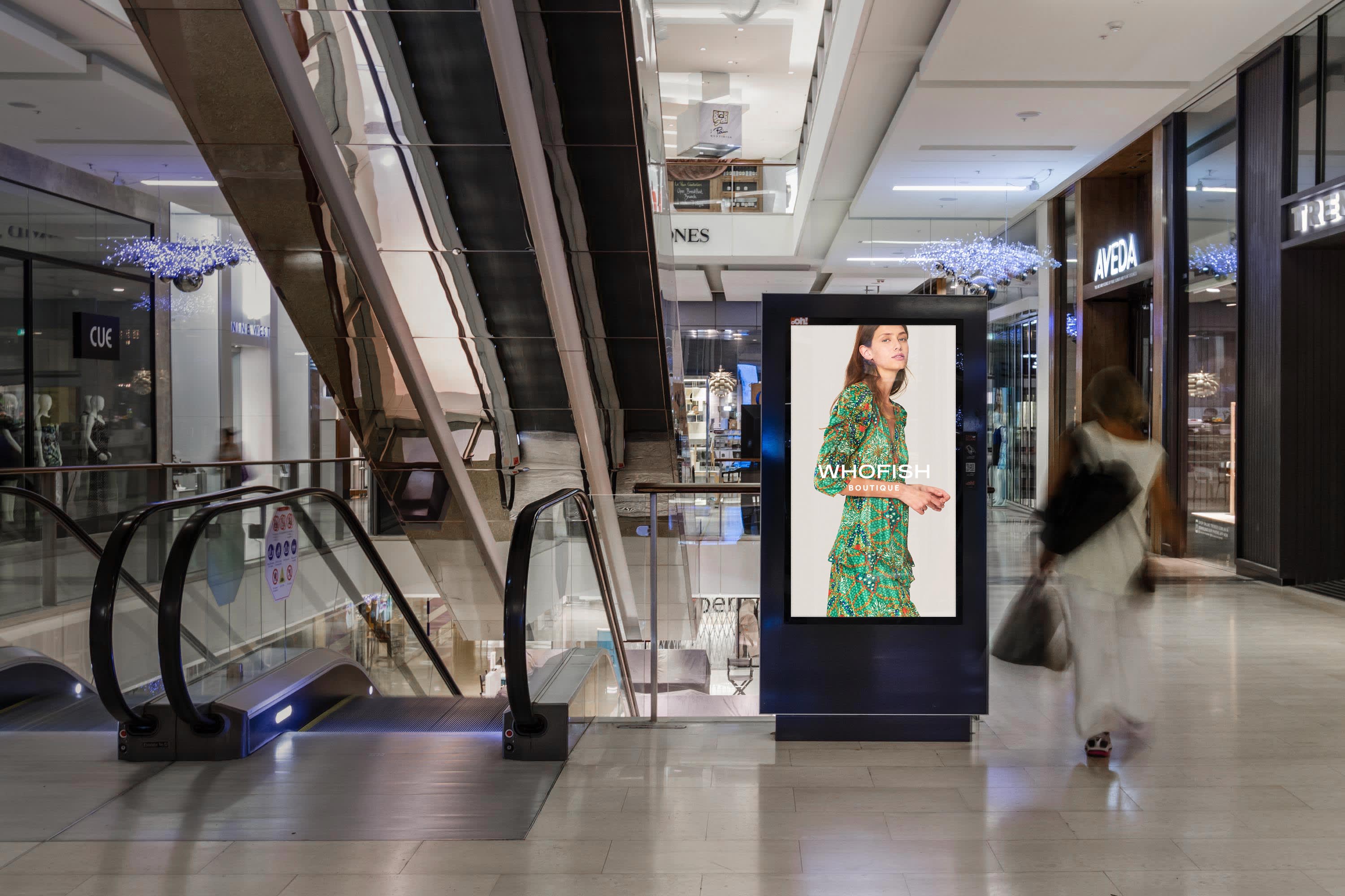 A shopping center. A woman walks past a digital billboard display showing a woman and the Whofish brand