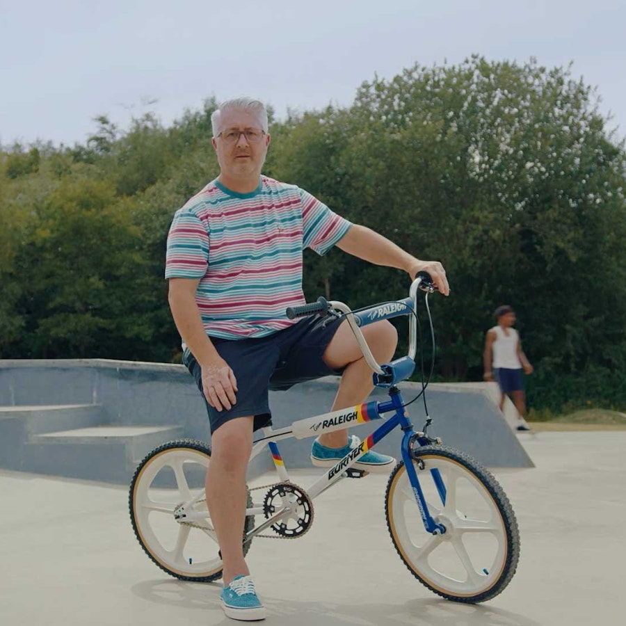 A grey haired man in a striped tshirt and shorts sat on a Raleigh BMX in a skate park