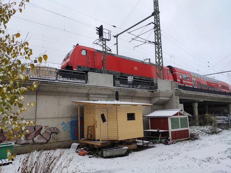 A sauna building located under train tracks, with a red train passing. There is snow on the ground.