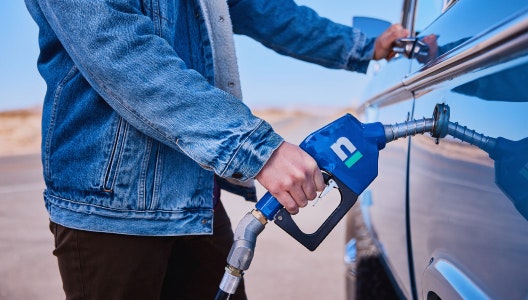 A photo of a person pumping gasoline into their car. The gasoline pump is blue with the Nacero logo.
