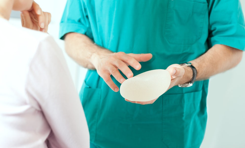 Doctor in green scrubs holding a breast implant and showing it to a female patient.