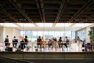 People sit on stools at high round tables in a light-filled room as musicians play.