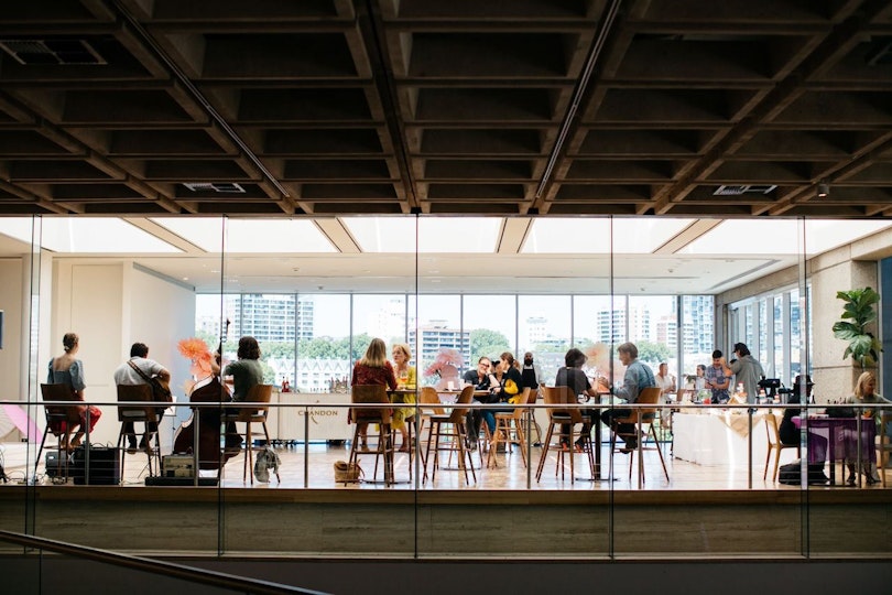 People sit on stools at high round tables in a light-filled room as musicians play.