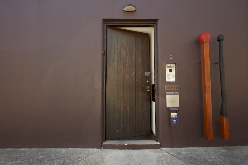 A wooden door slightly ajar with two large matchstick sculptures next to the doorbell.