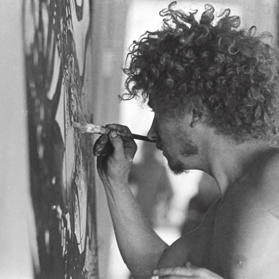 A profile view of a white man with short ringlets who is bare-chested. He is applying paint to a canvas using a paintbrush in his right hand.