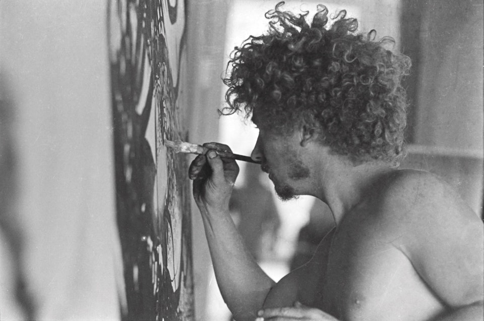 A profile view of a white man with short ringlets who is bare-chested. He is applying paint to a canvas using a paintbrush in his right hand.