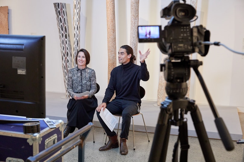Two people seated in front of an artwork. In the foreground is a camera and a monitor.