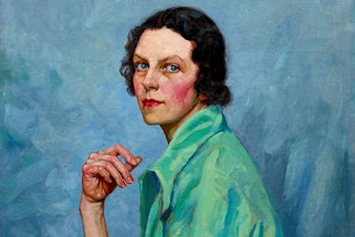A woman sits with one elbow resting on her thigh and her hand held in front of her chest. She wears a wide-collared, short-sleeved green shirt and has wavy, collar-length dark hair, blue eyes and pink cheeks and lips.