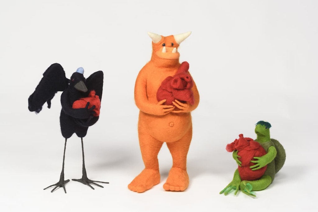 Three creatures made from stuffed felt, each holding a red object. From left, a standing black bird, a standing orange figure with horns from its head and snout, a seated green turtle.