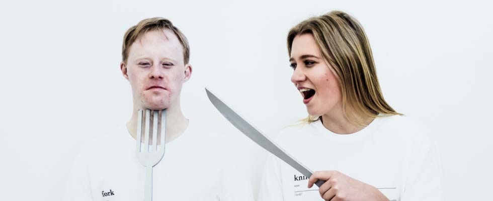 A person wearing a t-shirt with the word 'fork' holds the prongs of a giant fork under their chin while on their left a person wearing a t-shirt with the word knife holds a large knife-like object.