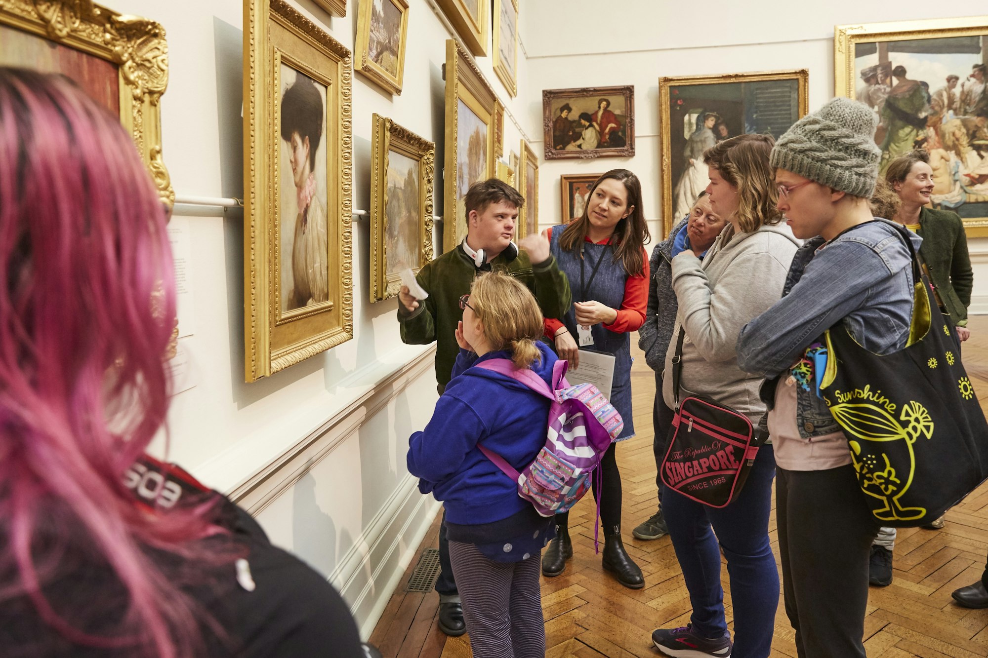 A person with short brown hair and light skin stands in front of a salon hang of paintings. There are 8 people around them.