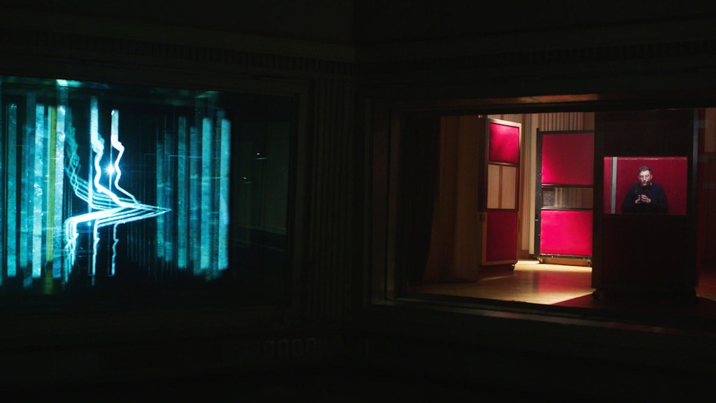 Video still of a dark room with vertical streams of blue-green light on the left and a room with red material and a video monitor on the right.