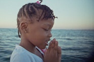 Video still of a child with clasped hands in front of the sea.