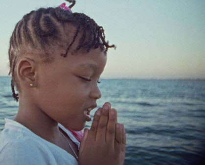 Video still of a child with clasped hands in front of the sea.