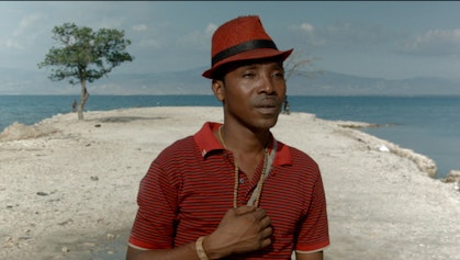 Video still of a person in a red hat and red polo shirt. In the background is the sea and a tree.