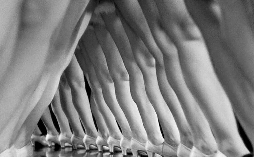 Video still in black and white of many legs with low-heeled shoes in an A shape seen from a low angle.