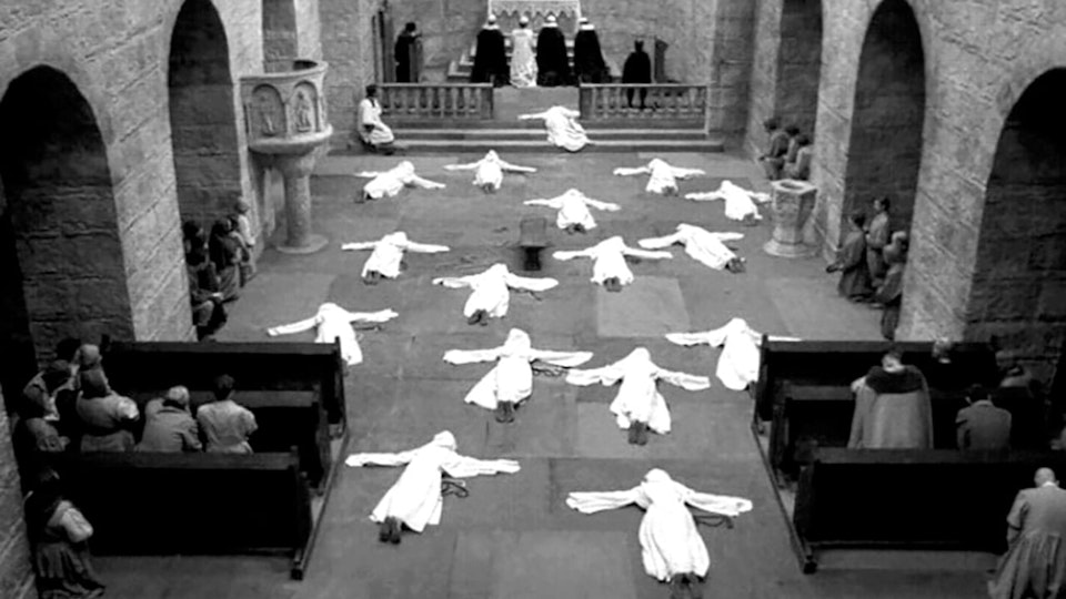 Video still in black and white of 16 nuns in white outfits lying face down on  the floor of a church.