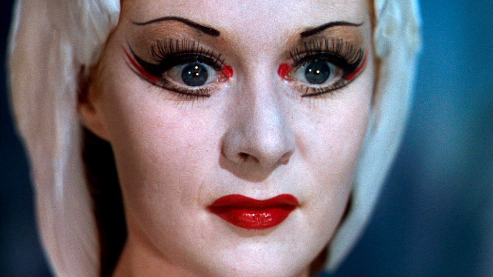 Video still of a close-up of a face with painted eyebrows, eye makeup and bright red lipstick.