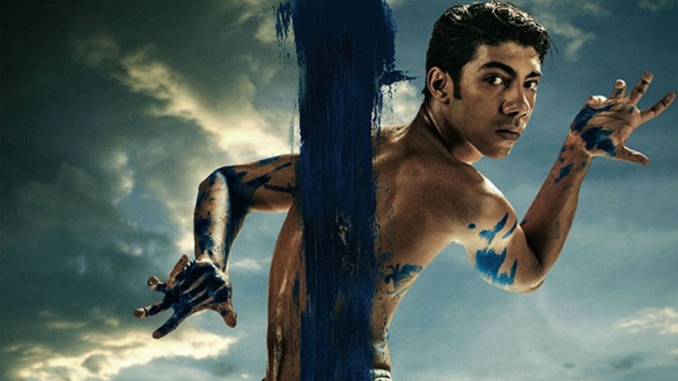 A person holding their arms in opposite angles in front a blue sky. There is blue paint down the middle of the image and splashed all over the torso.