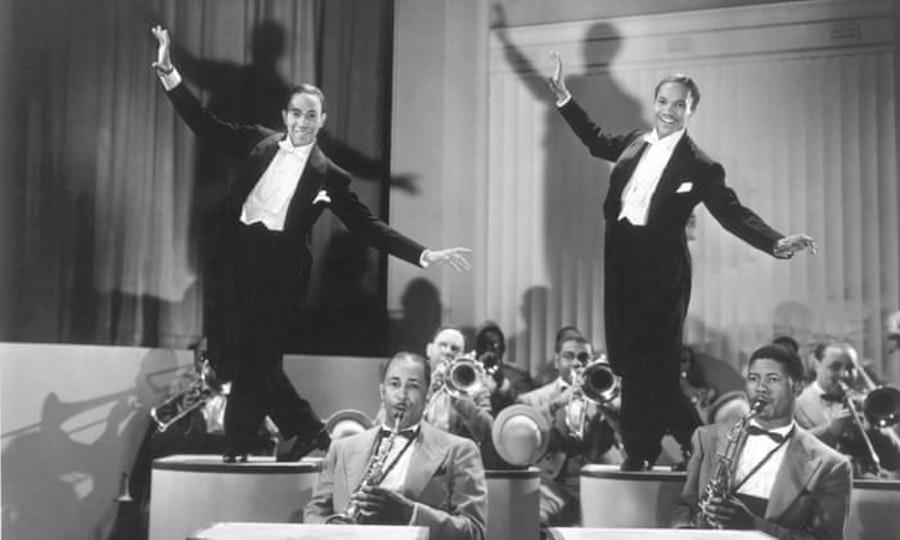Video still of two people in tuxedos dancing high up on podiums. A band plays beneath them.