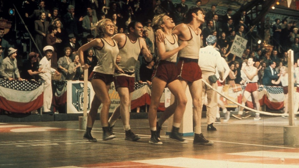 Video still of a group of four people running in gym clothing. In the background are spectators.