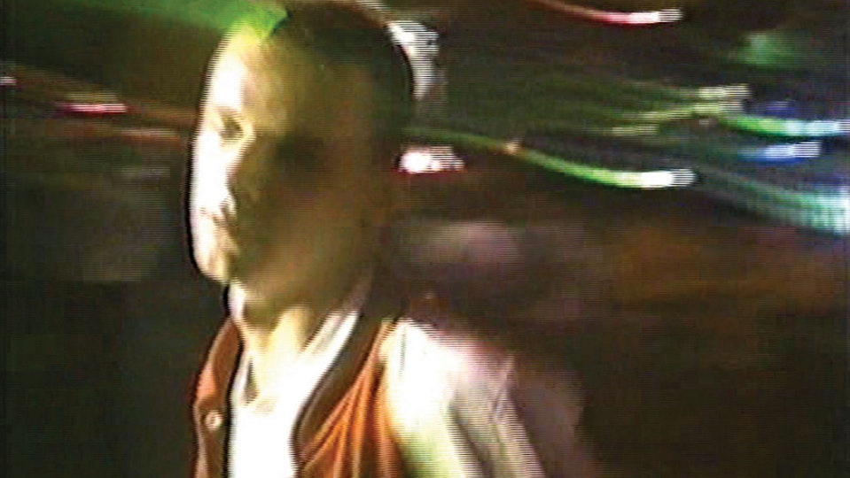 A young person with shaved hair dyed green in front of a blurry background of colourful lights.