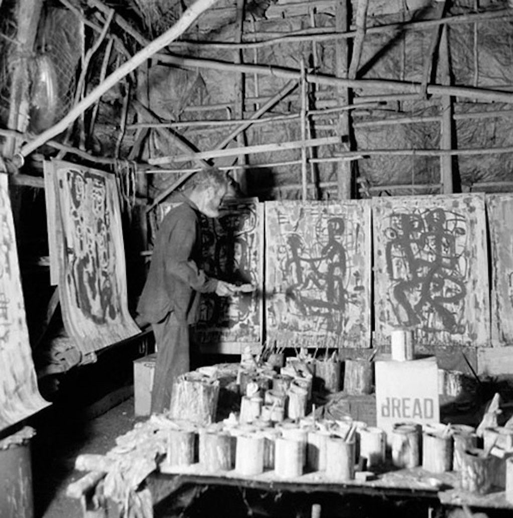A black-and-white photo of a person in a grass and wood building, working on one of several unframed paintings next to a table laden with paint tins.