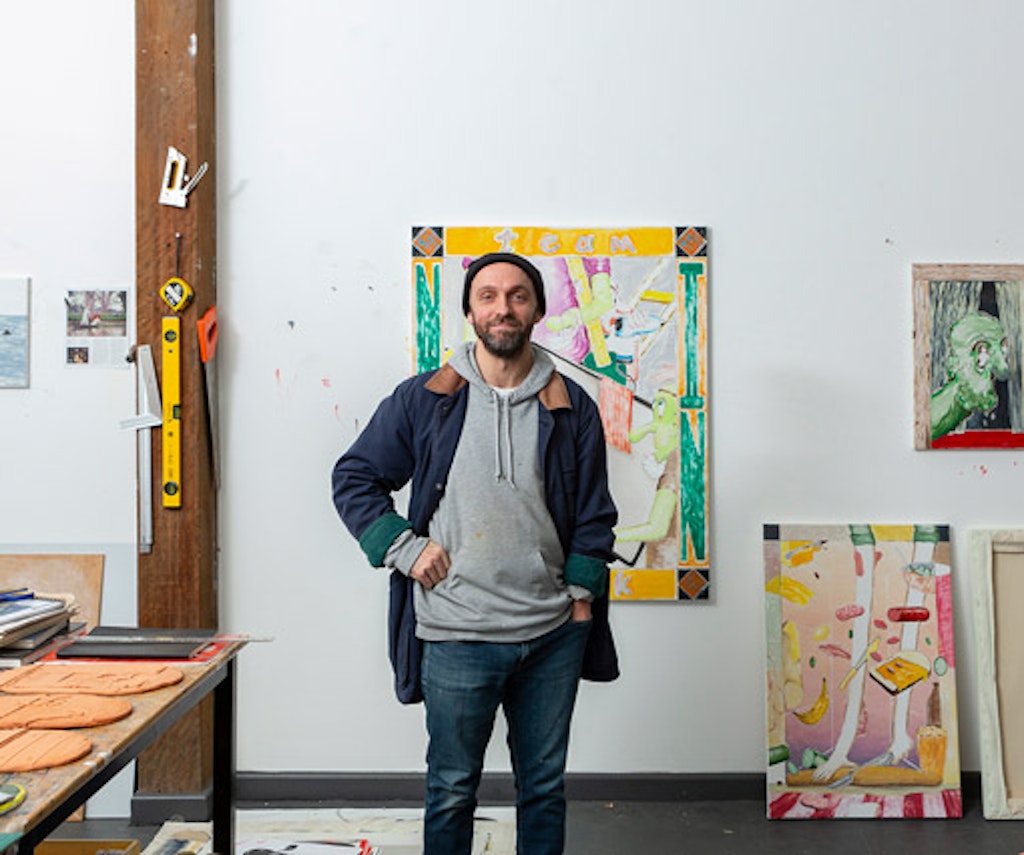 A person stands in a room with a few works of art on or against the wall behind them.