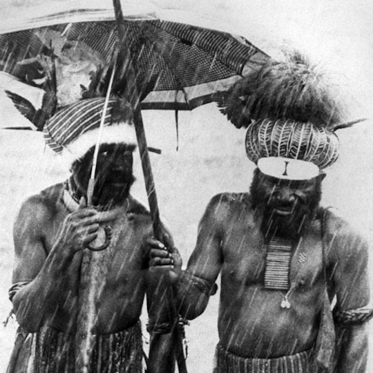 Two people with dark brown skin and wearing headdresses stand in the rain. One. person holds an umbrella over both of them.