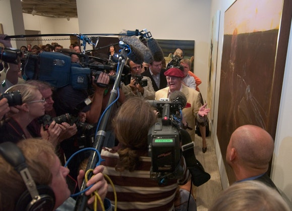 A person in a red beret stands in front of a crowd of people with TV cameras and microphones next to a painting on a wall