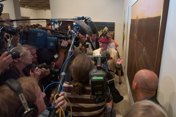 A person in a red beret stands in front of a crowd of people with TV cameras and microphones next to a painting on a wall