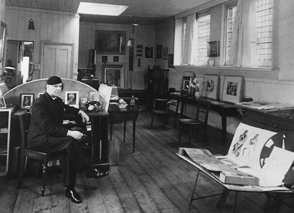 A black-and-white photo of a person sitting at a desk in a room in which artworks lie or sit propped on tables.