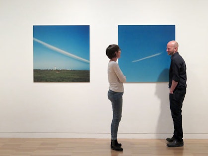 Two people in conversation in front of two square artworks on a gallery wall.