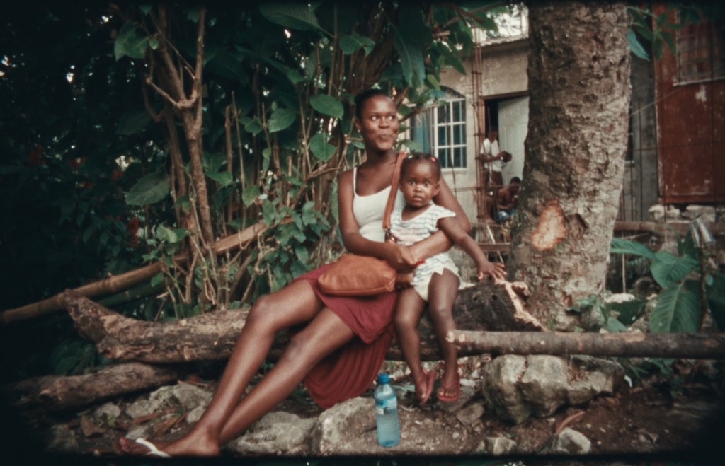 A person with medium-dark skin tone and closely cropped black hair wearing a single and a skirt. They are sitting on a fallen tree and holding a toddler close to them.