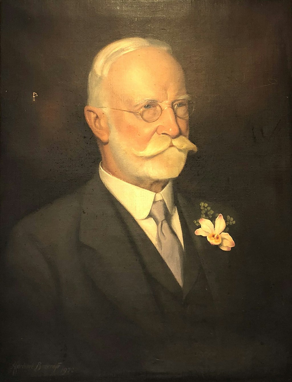 White-haired man in wire-framed round glasses, with a neatly trimmed beard and luxurious moustache wearing a three-piece suit and tie, with an orchid on his lapel