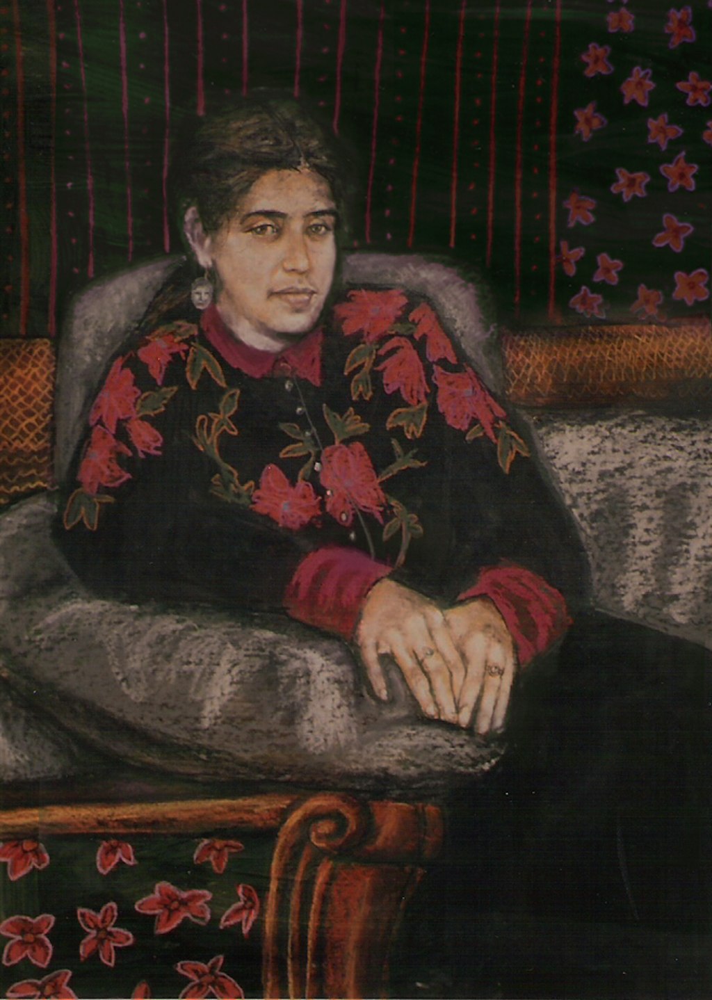 Woman in a grey armchair, her two hands on its right arm. Her long dark hair is pulled back, revealing an earring with a face dangling from her right ear. She is wearing dark buttoned-up top with dark pink cuffs, collar and flowers and dark green leaves. A similar floral motif is in background wallpaper and foreground furniture.